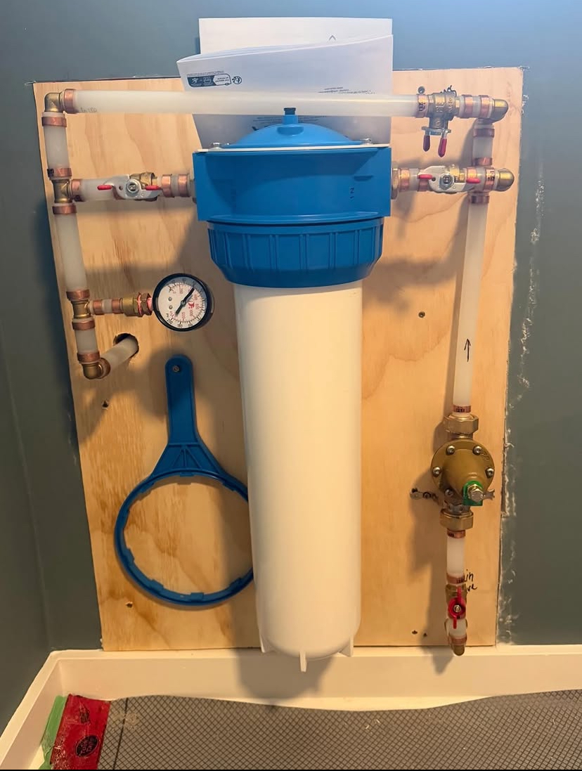 Whole-house water filter mounted on plywood with copper piping, shutoff valves and a pressure gauge
